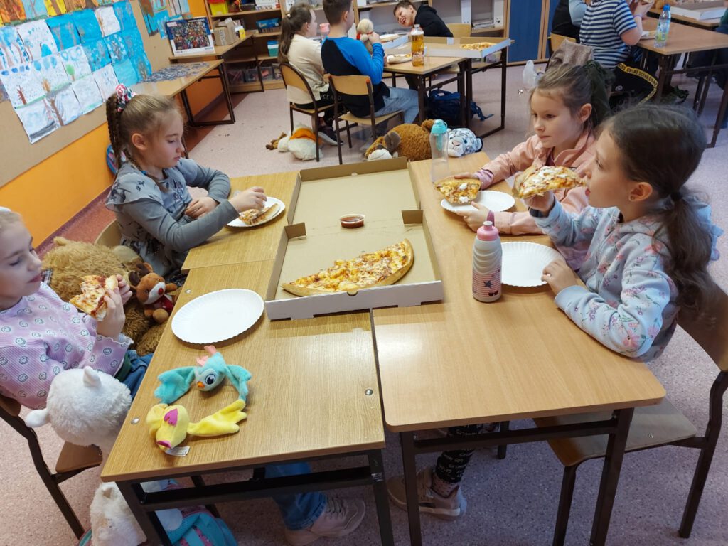 Na zdjęciu uczniowie klasy drugiej A siedzą przy stolikach i jedzą pizzę. 