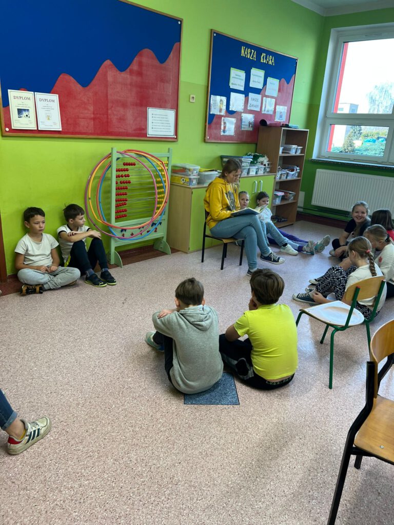 Uczennica klasy 8  siedzi na krzesełku, wokół niej siedzą uczniowie klasy 3b i słuchają, jak starsza koleżanka czyta im książkę w ramach akcji "Cała Polska czyta dzieciom".