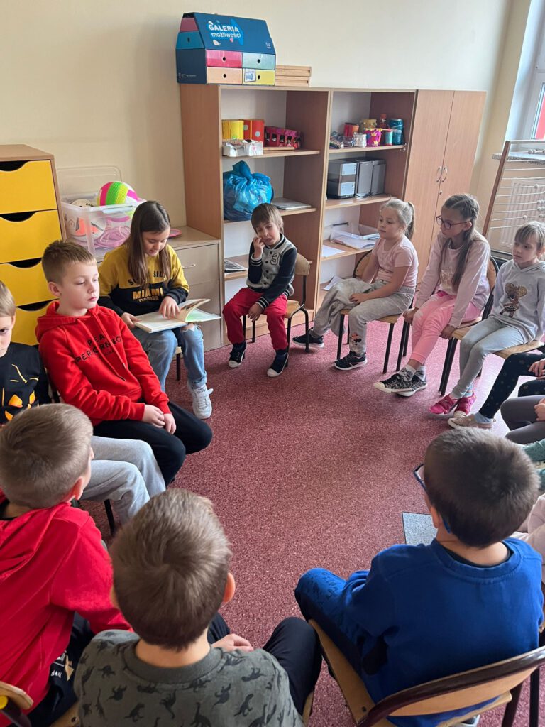 Uczennica klasy 4  siedzi na krzesełku, wokół niej siedzą uczniowie klasy 1b i słuchają, jak starsza koleżanka czyta im książkę w ramach akcji "Cała Polska czyta dzieciom".