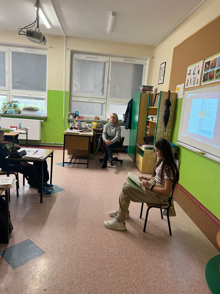 Uczennica klasy 7 siedzi na krzesełku, przed nią siedzą uczniowie klasy 3a i słuchają, jak starsza koleżanka czyta im książkę w ramach akcji "Cała Polska czyta dzieciom".