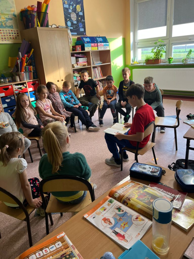 Uczeń klasy 4  siedzi na krzesełku, wokół niego siedzą uczniowie klasy 1 a i słuchają, jak starszy kolega czyta im książkę w ramach akcji "Cała Polska czyta dzieciom".