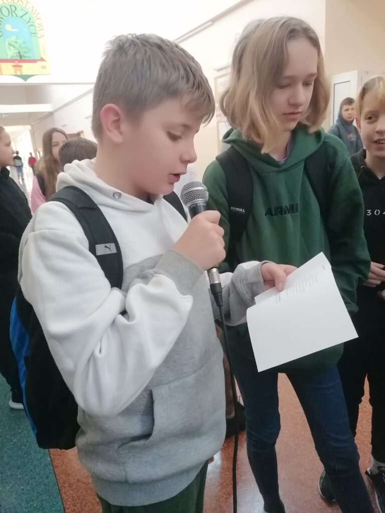 Uczeń trzyma mikrofon i kartkę, na której znajduje się jeden z łamańców językowych, próbuje z poprawna wymową przeczytać go na głos. 