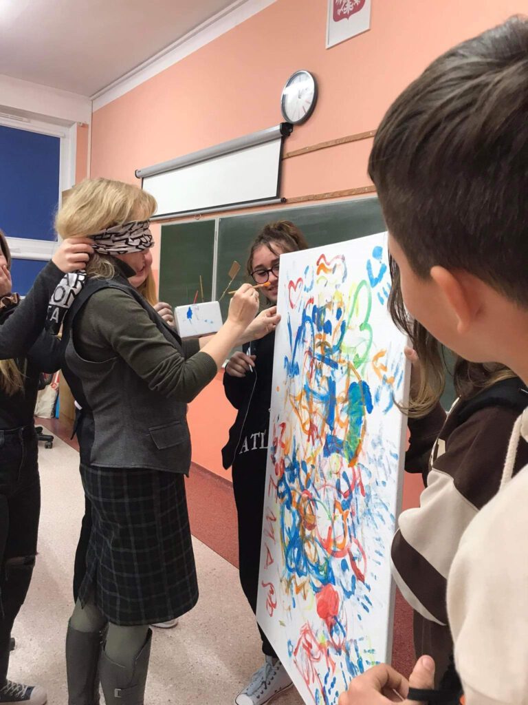  Jedna z klas. Nauczycielka stoi z zasłoniętymi oczami i maluje obraz. Z lewej strony stoją uczniowie, którzy trzymają płótno do malowania.