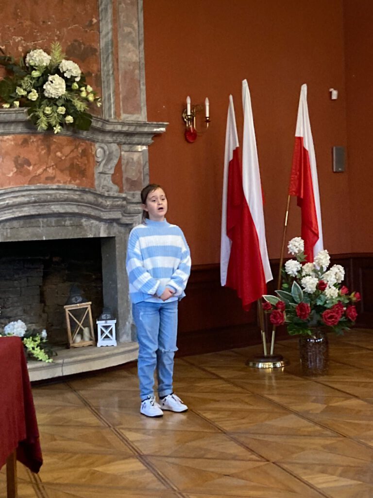 Na zdjęciu stoi uczennica i recytuje wiersz w XVI Patriotycznym Powiatowym Konkurs Recytatorskim  pod nazwą „Pokolenie” (kategoria klasy IV-V).