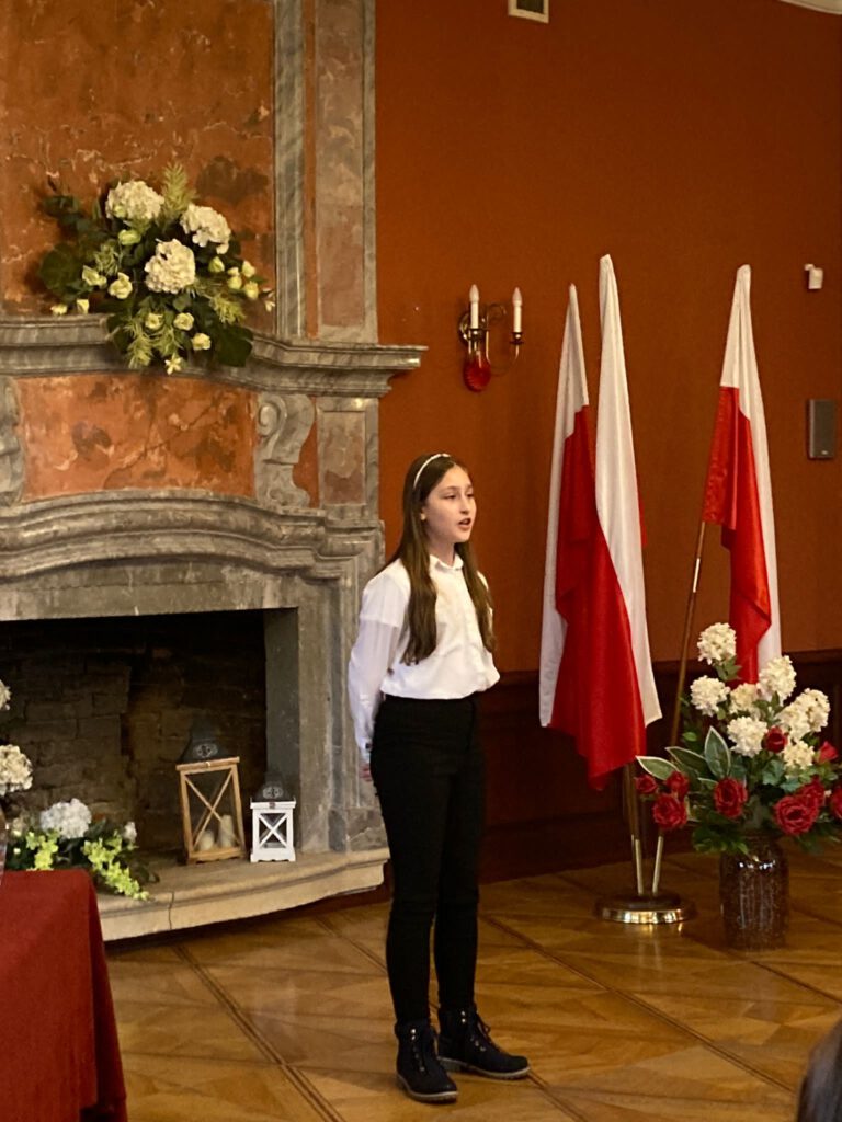 Na zdjęciu stoi uczennica, ubrana w strój galowy  i recytuje wiersz w XVI Patriotycznym Powiatowym Konkurs Recytatorskim  pod nazwą „Pokolenie” (kategoria klasy VII-VIII). 