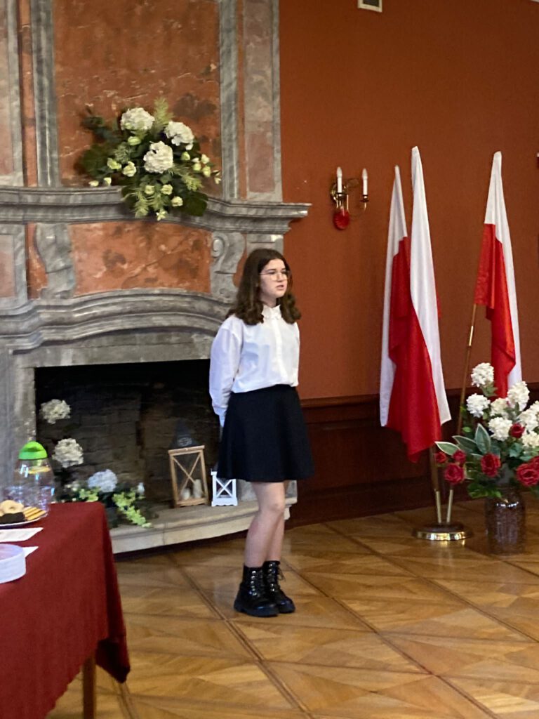 Na zdjęciu stoi uczennica, ubrana w strój galowy  i recytuje wiersz w XVI Patriotycznym Powiatowym Konkurs Recytatorskim  pod nazwą „Pokolenie” (kategoria klasy VII-VIII)