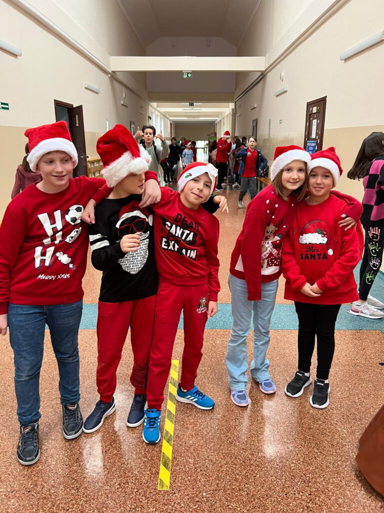 Zdjęcie prezentuje trzech chłopców i trzy dziewczynki z klasy 4, wszyscy ubrani są na czerwono i maja na głowie mikołajowe czapki. Uczniowie znajdują  się na szkolnym korytarzu, w tle widać innych uczniów. 