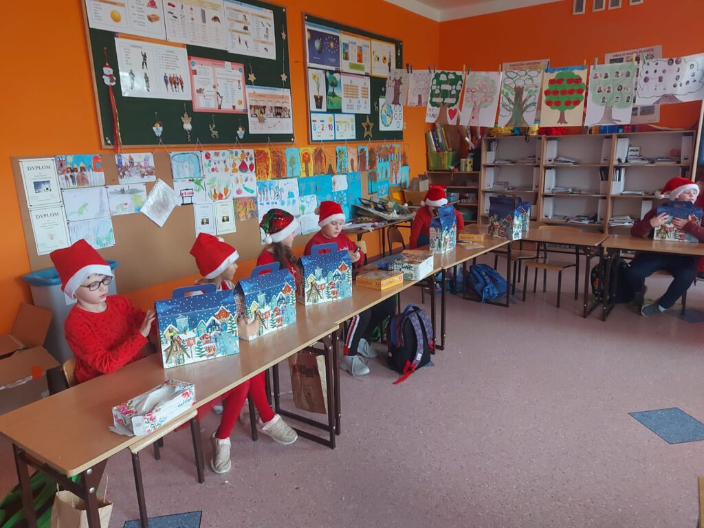 Zdjęcie przedstawia uczniów klasy 2a, uczniowie znajdują się w klasie, ubrani są na czerwono, na głowie mają mikołajowe czapki. Uczniowie siedzą w ławkach i oglądają otrzymany prezent. 