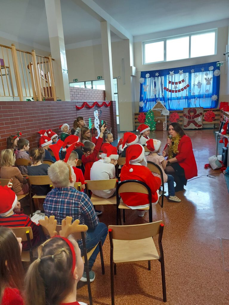 Na zdjęciu widać  uczniów ubranych świątecznie, nauczycielka zachęca uczniów o złożenia życzeń. podając mikrofon dzieciom.  