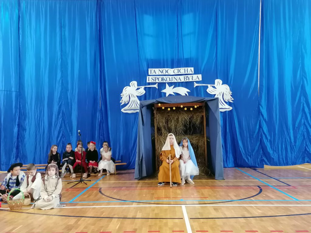 Na obrazku widać dzieci biorące udział w jasełkach: na środku przed szopką siedzi Józef z Maryją, po lewej stronie widać pasterzy i dzieci. 