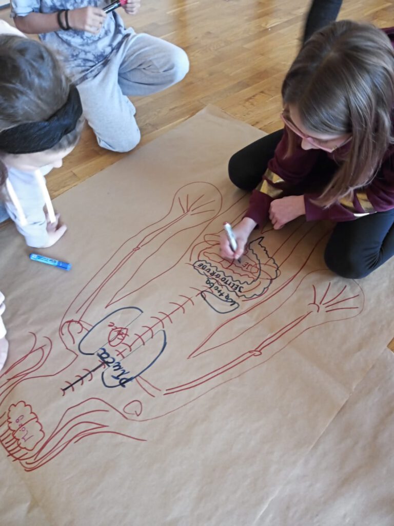 Na zdjęciu widać sześcioro dzieci, które na dużej kartce szarego papieru mają narysowane schemat postaci człowieka i zaznaczają organy ludzkie, które mają wpływa na wady postawy.  