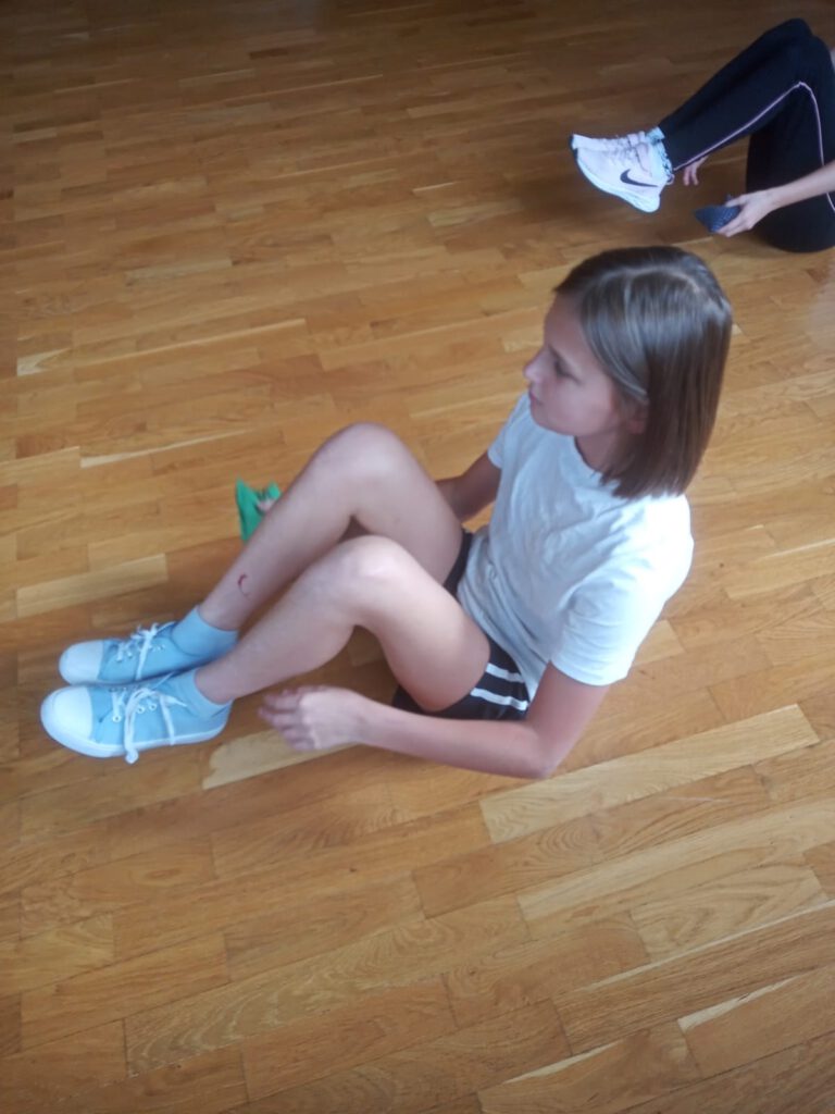 Na zdjęciu widoczna jest dziewczynka, które siedzi na  podłodze z nogami ugiętymi i przekłada pod kolanami woreczek gimnastyczny. Ubrana jest w białą koszulkę i ciemne spodenki.