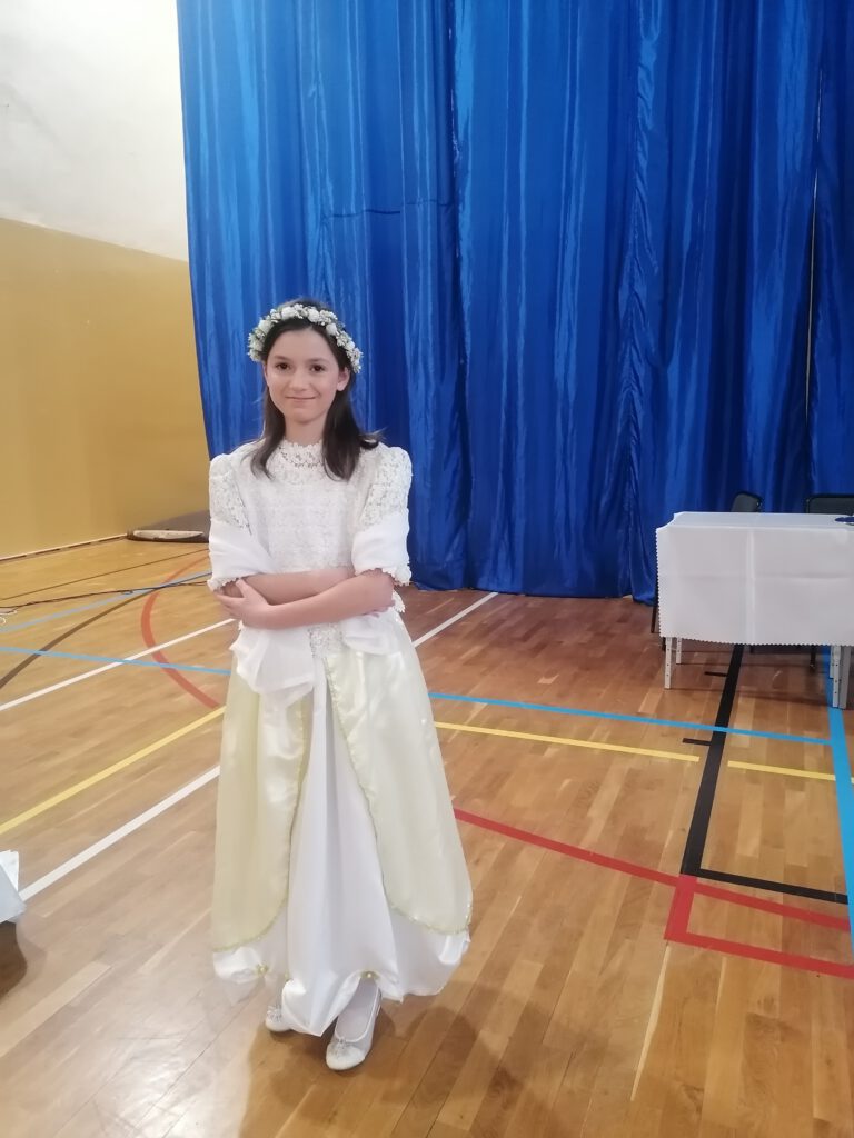 Zdjęcie przedstawia dziewczynkę ubraną w piękną białą sukienkę. Dziewczynka grała głowną role w przedstawieniu szkolnym "Kopciuszek".  