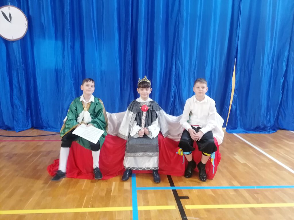Na zdjęciu trzech chłopców ubranych w piękne i wytworne stroje siedzi na krzesłach, uczniowie biorą udział w przedstawieniu szkolnym "Kopciuszek". 