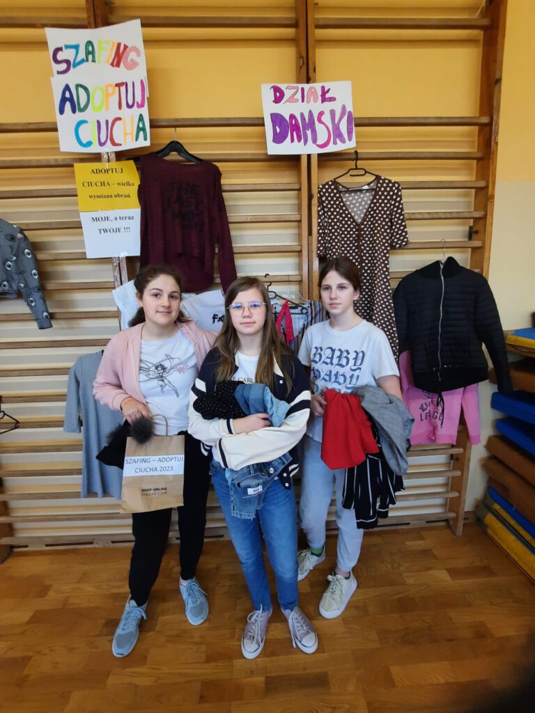 Na zdjęciu trzy uśmiechnięte dziewczynki trzymające w rękach ubrania pochodzące z wielkiej wymiany ubrań. Nad nimi plakat z hasłem: Szafing - Adoptuj ciucha!