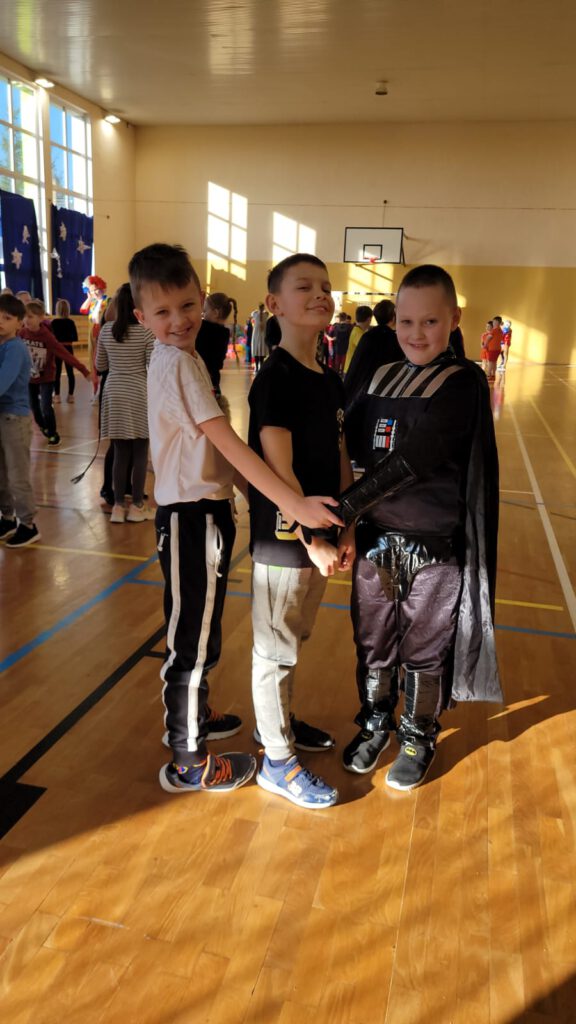 Zdjęcie przedstawia trzech chłopców, dwóch z nich ma na sobie strój sportowy, trzeci chłopiec ma na sobie strój z filmu "Gwiezdne wojny".  Chłopcy są uśmiechnięci, w tle inne bawiące się dzieci. 