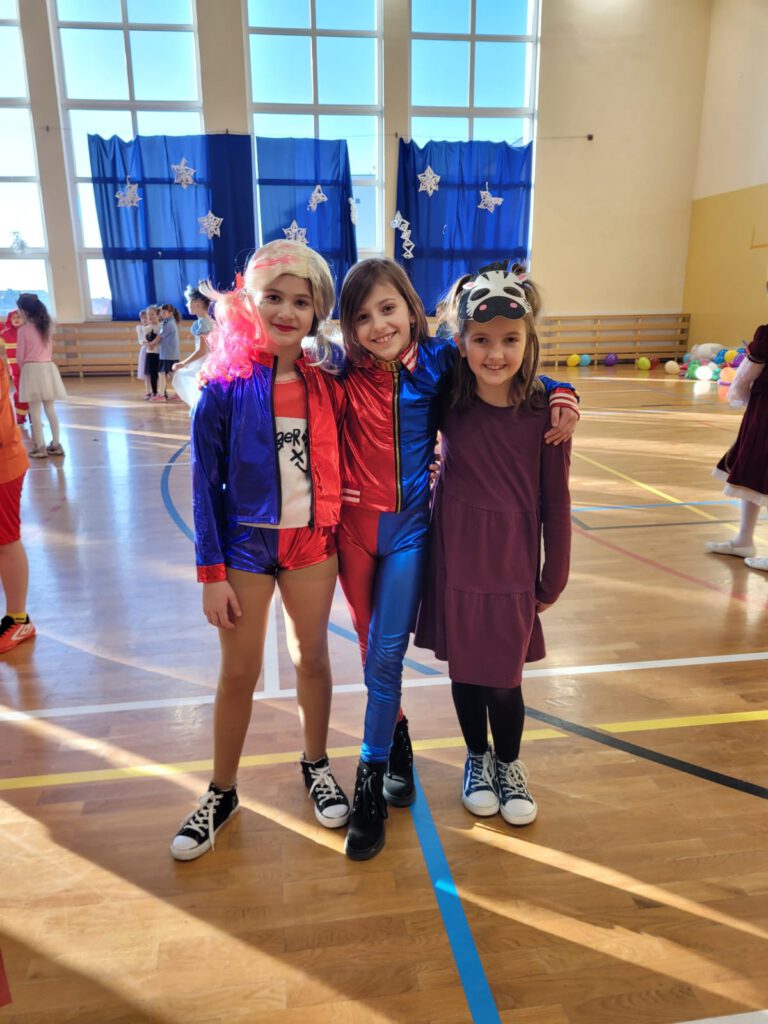 Zdjęcie przedstawia trzy dziewczynki ubrane w kolorowe stroje, dwie maja na sobie strój niebiesko-czerwony a trzecia dziewczynka ma na głowie maskę zebry. Dziewczynki sa bardzo uśmiechnięte.  