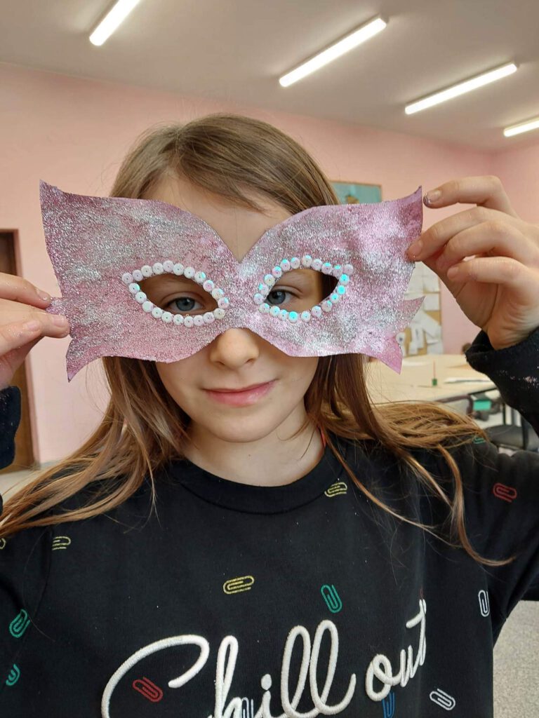 Przygotowania do balu karnawałowego- maski karnawałowe. Na zdjęciu dziewczynka ma na twarzy maskę fioletową i posypaną srebrnym brokatem, wokół oczy ma srebrne cekiny. 