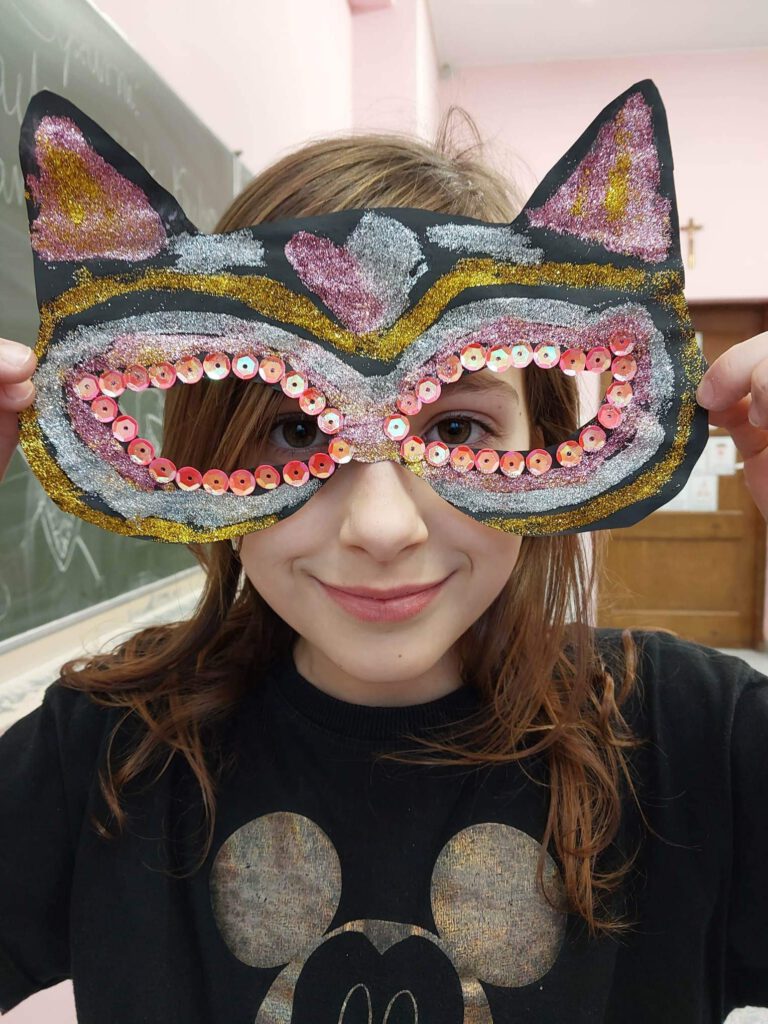 Przygotowania do balu karnawałowego- maski karnawałowe. Na zdjęciu dziewczynka ma na twarzy maskę karnawałową w kształcie kota, maska jest czarno-złoto-srebrna i posypana złotym i srebrnym brokatem. 