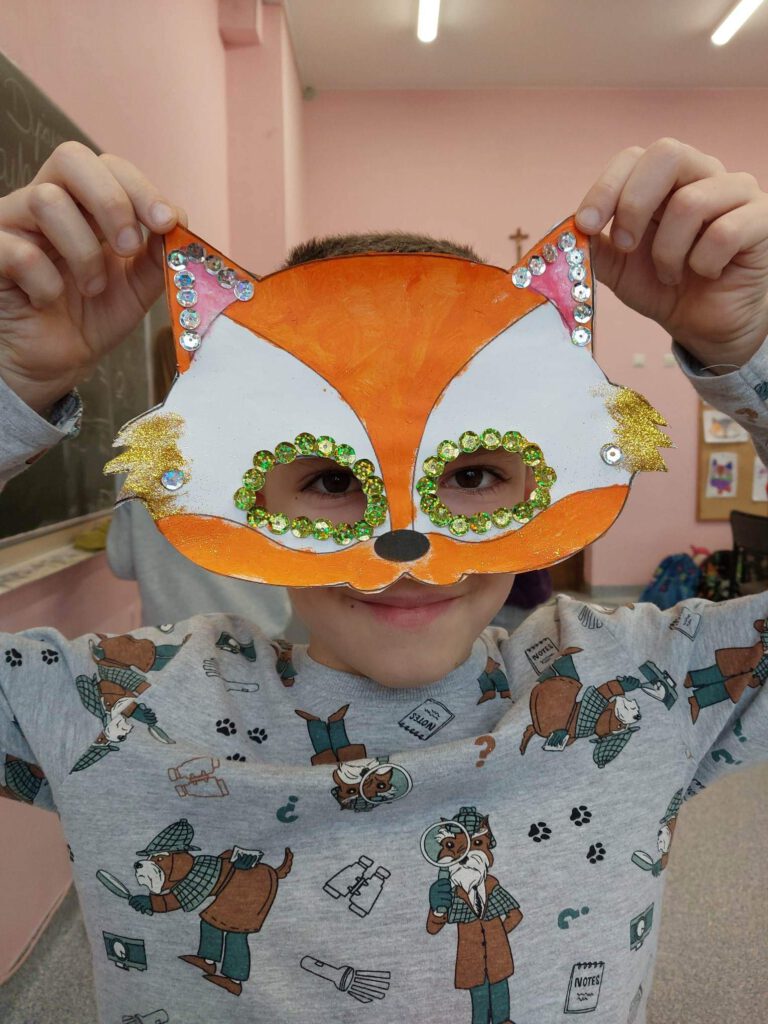 Przygotowania do balu karnawałowego- maski karnawałowe. Na zdjęciu chłopiec ma na twarzy maskę karnawałową w kształcie  lisa, maska jest biało-pomarańczowa i na uszach oraz wokół oczu ma srebrne i złote cekiny. 