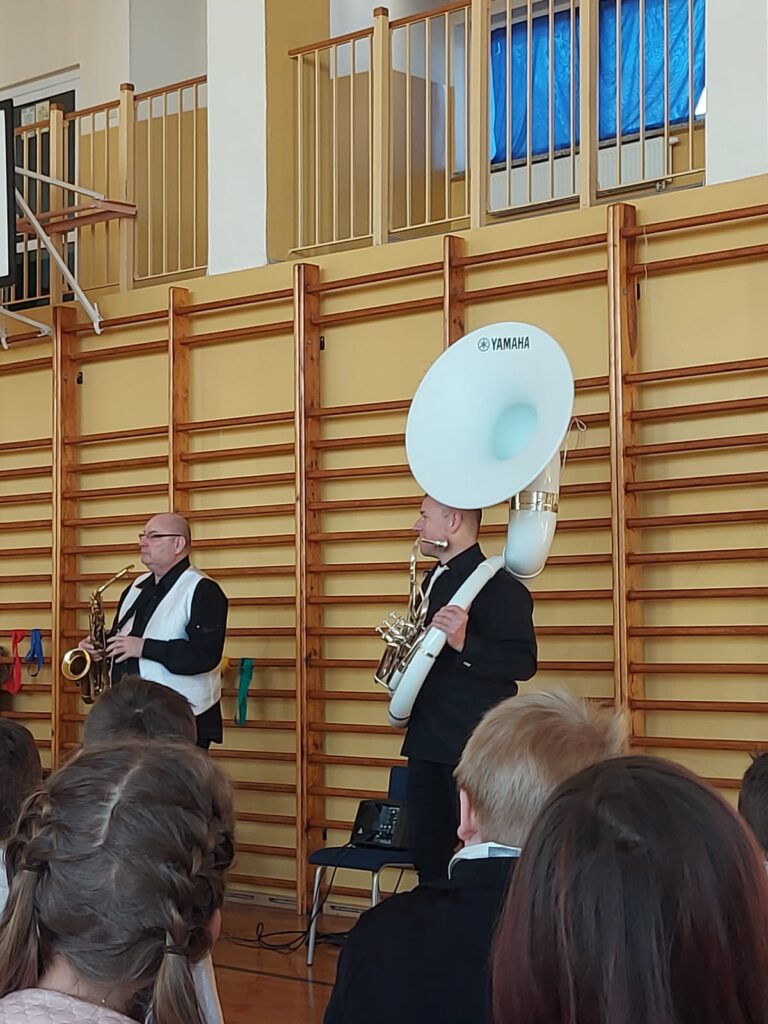 Zdjęcie  przedstawia muzyków, jeden z nich trzyma saksofon, drugi trzyma suzafon. 