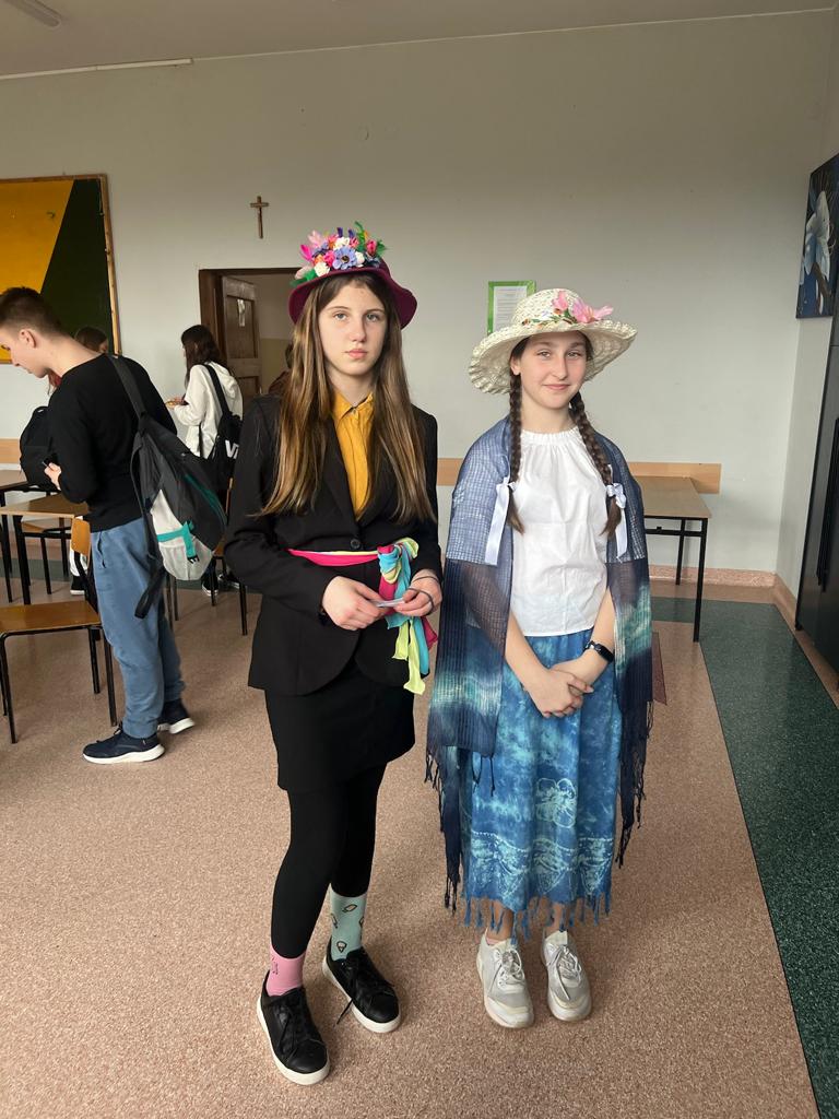 Na zdjęciu stoją dwie dziewczynki, na głowie  mają kapelusze z kwiatkami. Jedna z dziewczynek ma na ramionach chustę koloru grafitowego , druga w pasie ma związany kolorowy materiałowy pasek. 