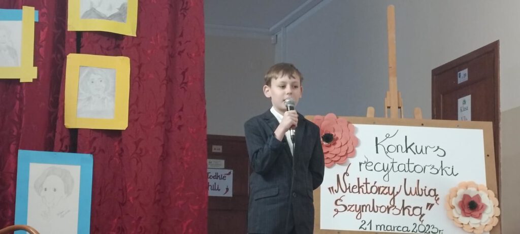 Zdjęcie przedstawia ucznia, który stoi obok plakaty z napisem "Konkurs recytatorski". Uczeń trzyma mikrofon i recytuje wiersz. 
