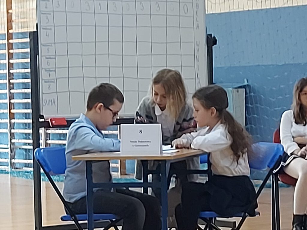 Powiatowy Turniej Wiedzy Klas II w Polkowicach- troje uczniów siedzi przy stoliku i rozwiązuje  zadania konkursowe. 