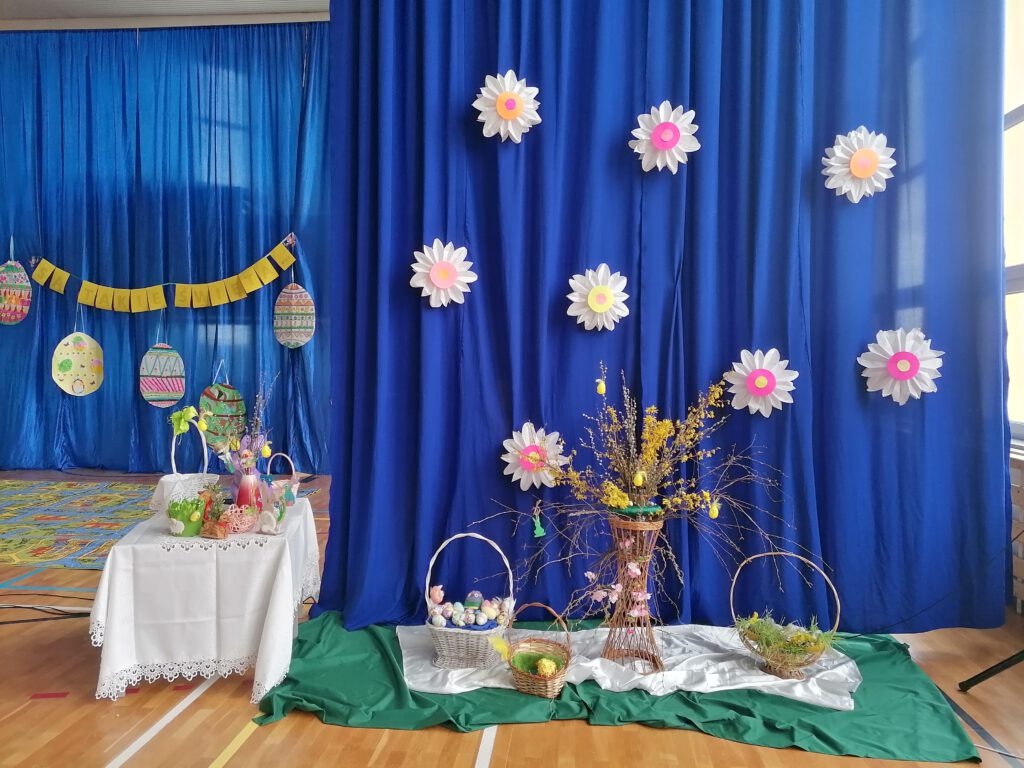 Apel Wielkanocny- dekoracja sali. Na zdjęciu widać stół z białym obrusem na podłodze stoją trzy koszyki, w tle kwiatki wykonane z paieru.