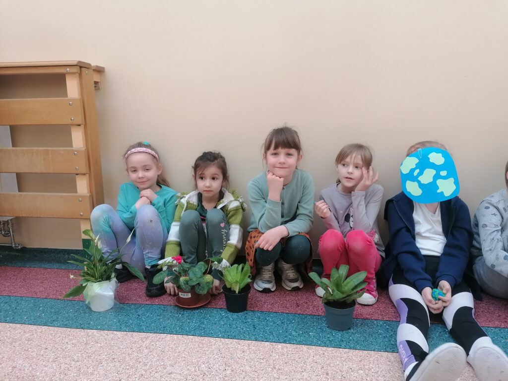 Na zdjęciu widać pięć dziewczynek z klasy pierwszej, przed nimi stoją na podłodze kwiatki w doniczkach. 