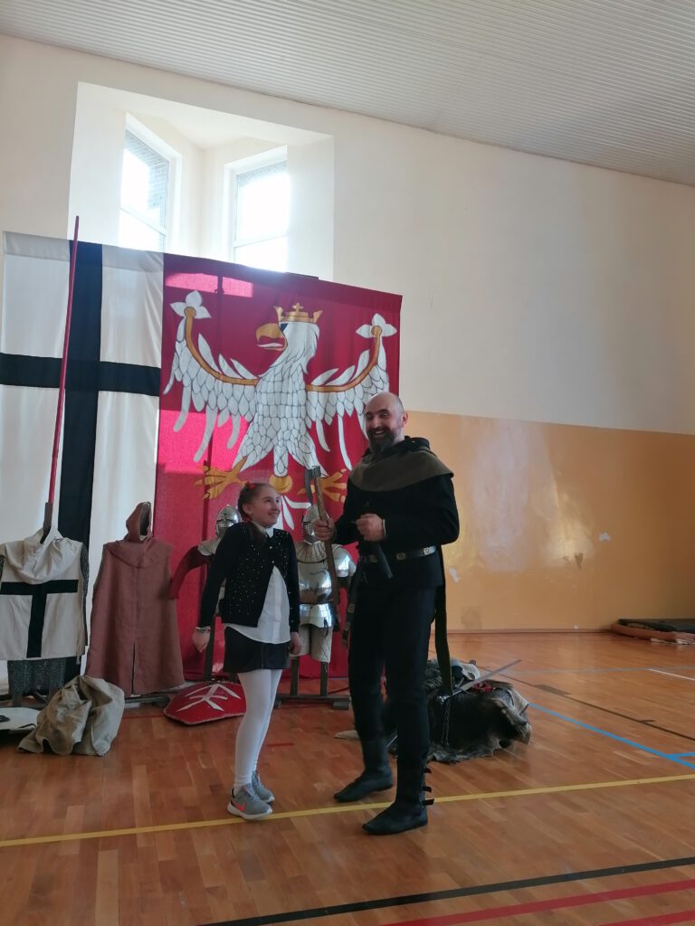 Żywa lekcja historii "Na Grunwald"- na zdjęciu stoi dziewczynka, obok niej prowadzący Żywą lekcję historii, ubrany jest w strój ze średniowiecza, w ręce trzyma topór. 