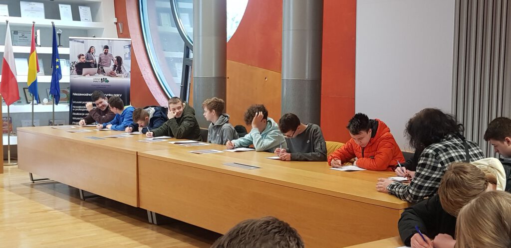 Uczniowie uczestniczą w spotkaniu z doradcą zawodowym w siedzibie Starostwa Powiatowego w Polkowicach. W tle widać flagi Polski oraz Unii Europejskiej. Za uczniami znajduje się duże okrągłe okno.