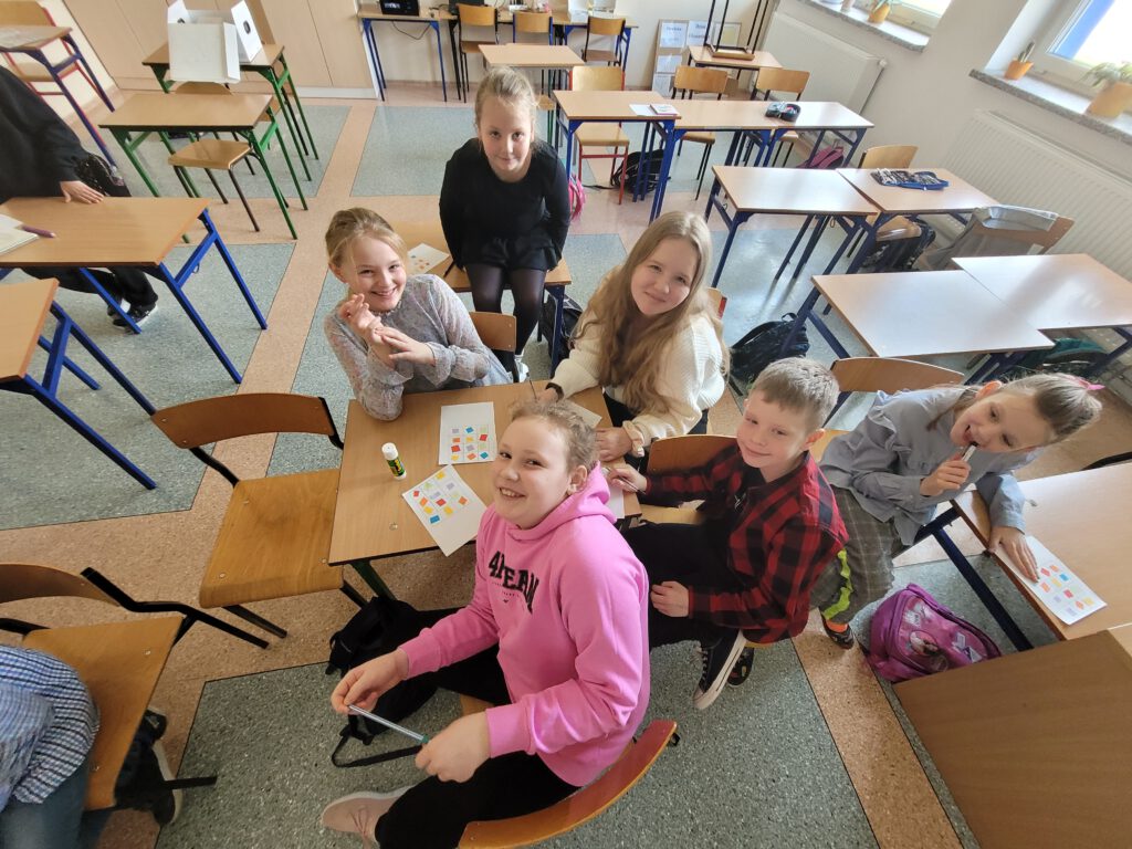 Laboratoria Przyszłości- Gra w Bingo z Robotem Photon. Zdęcie przedstawia dzieci z klasy trzeciej, które siedzą przy stoliku i grają w bingo z robotem Photon. 