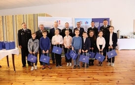 Powiatowe eliminacje Ogólnopolskiego  Turnieju Wiedzy Pożarniczej „Młodzież Zapobiega Pożarom”. Na zdjęciu stoją uczniowie, w ręce trzymają niebieskie torby 
 z nagrodami, za nimi stoją panowie ubrani w wyjściowe mundury strażackie. 