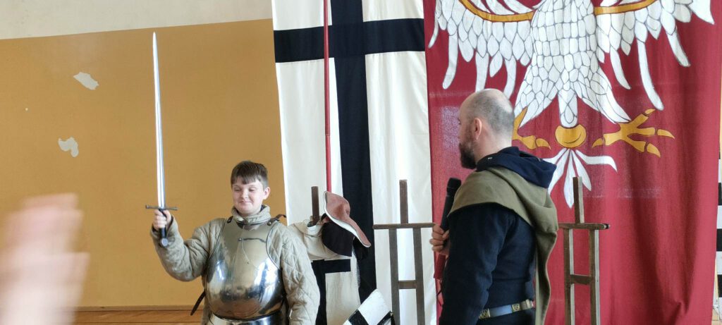 Żywa lekcja historii "Na Grunwald"- na zdjęciu chłopiec trzyma w prawej ręce  miecz, ubrany jest w zbroję rycerską, obok niego stoi p[prowadzący Żywą lekcję historii, w ręce trzyma mikrofon.  