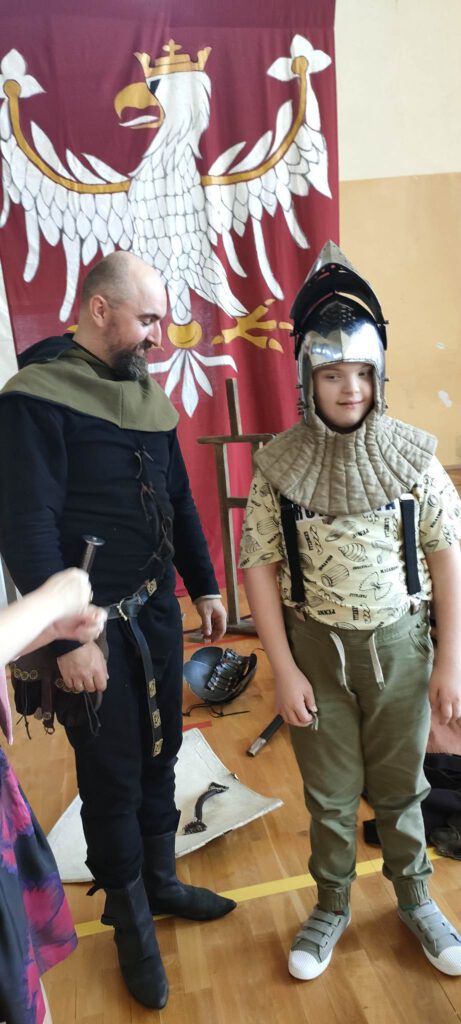Żywa lekcja historii "Na Grunwald"- na zdjęciu prowadzący Żywa lekcję historii, ubrany jest w strój ze średniowiecza,   obok niego stoi uczeń, na głowie ma hełm rycerski. 