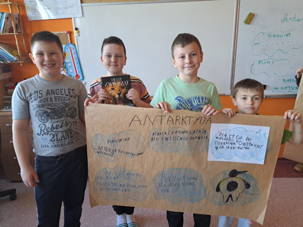Innowacja pedagogiczną "Lekturki spod chmurki-Podróże po świecie". Uczniowie prezentują  swój plakat o Antarktydzie. Stojąc,  trzymają szary papier na którym narysowali niebieskie chmurki z informacjami o Antarktydzie. Buzie chłopców są uśmiechnięte. 