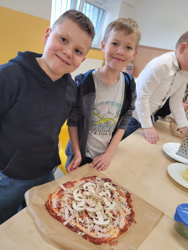 Pizza na zajęciach świetlicowych. Na zdjęciu dwóch uśmiechniętych chłopców prezentuje swoją pizzę z dodatkami. 