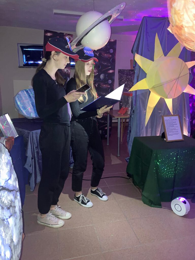 Szkolne obchody roku Mikołaja Kopernika. Nas zdjęciu uczennice klasy 6 b czytają opisy planet. Nad głowami wiszą modele planet. 