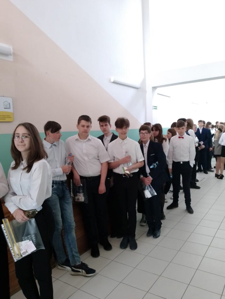 Egzamin ósmoklasisty. Na zdjęciu uczniowie klasy 8 a ubrani na galowo  czekają przed salę gimnastyczną, na losowanie numerków stolików. 