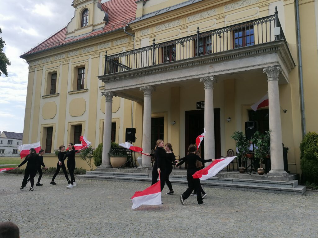 Obchody Konstytucji 3-go maja i świąt majowych. Na zdjęciu sześć dziewczynek ubranych na czarno wykonuje taniec  z flagami biało-czerwonymi. 