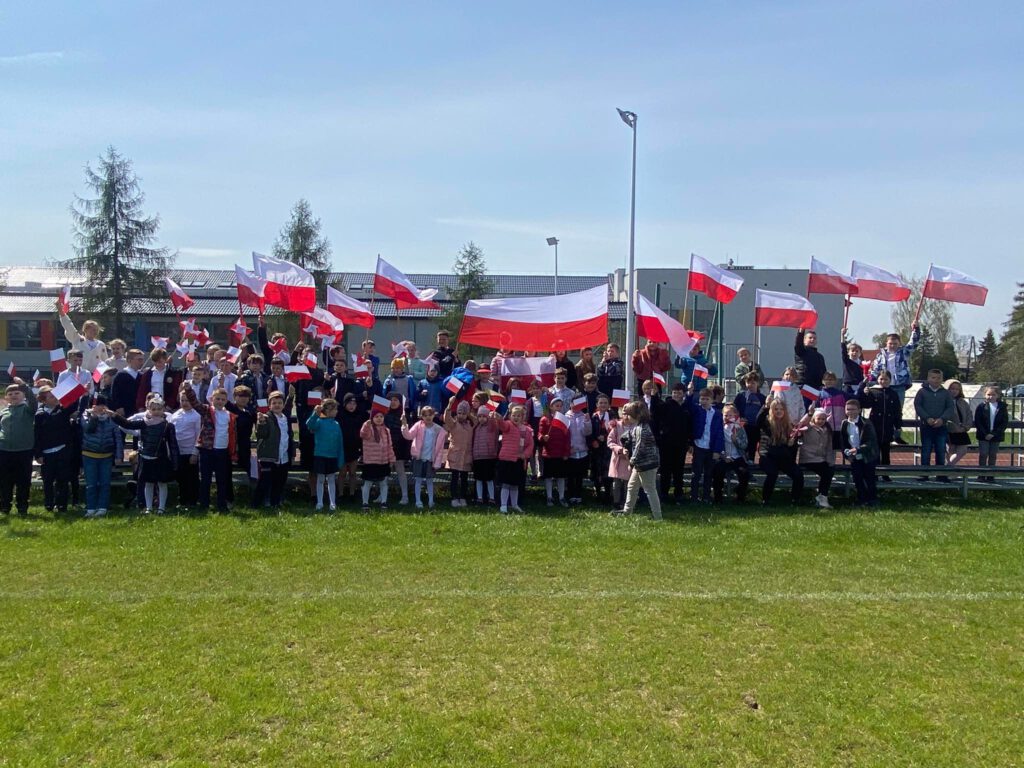 Spacer patriotyczny z okazji świat majowych. Na zdjęciu uczniowie pozują do zdjęcia grupowego, wznoszą do góry swoje flagi i chorągiewki biało-czerwone. 