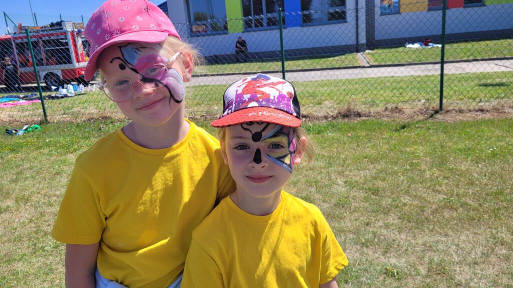 Międzynarodowy Dzień Dziecka na sportowo, zabawnie i kolorowo- „smaki z dzieciństwa-smaki świata- Chile”. Na zdjęciu uczennice klasy 1 a, w żółtych koszulkach i czapeczkach z daszkiem. Po lewej stronie twarzy mają namalowane połowę kolorowego motyla. 