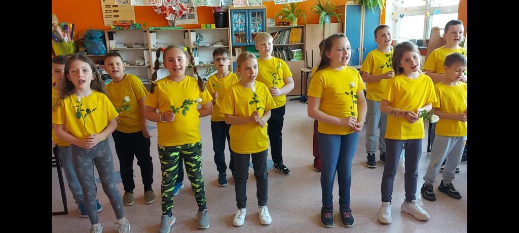 Klasa 2 a - Dzień Matki. Zdjęcie przedstawia uczniów klasy 2a, ubranych w żółte koszulki i śpiewających  piosenkę. 