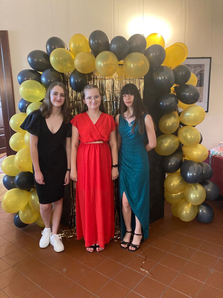 Bal ósmoklasistów 2023. Na zdjęciu trzy dziewczynki klas ósmych stoją na tle dekoracji z czarnymi i złotymi balonami.  