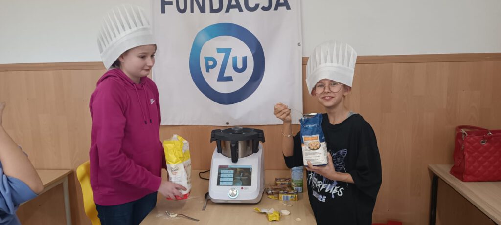 Podsumowanie projektu "Z Fundacją PZU po lekcjach". Na zdjęciu uczennice w białych kucharskich czapkach podczas warsztatów kulinarnych. 