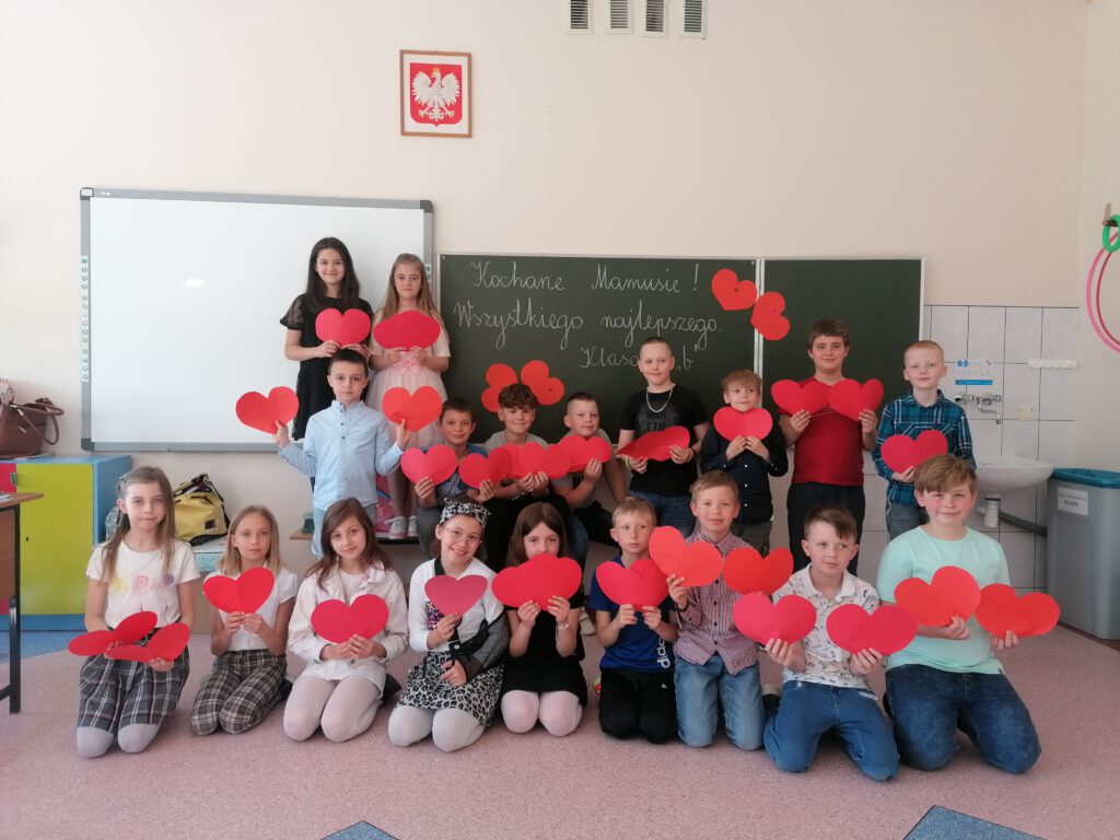 Klasa 2 b - Dzień Matki. Uczniowie klasy 2 b ustawieni na tle tablicy szkolnej pozują do zdjęcia trzymając w rękach czerwone serca. 