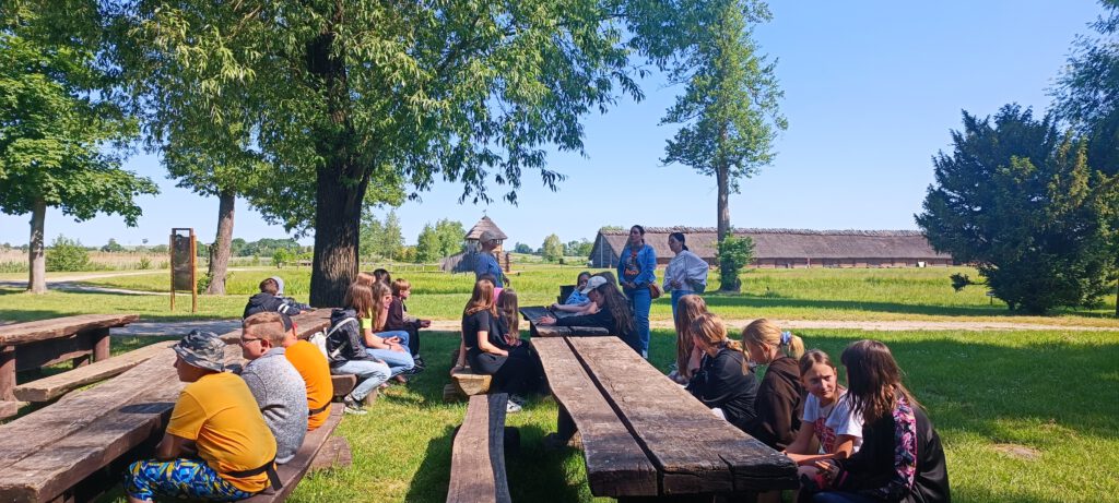 Dwudniowa wycieczka do Biskupina i Torunia w ramach programu "Poznaj Polskę".  Uczniowie siedzą na drewnianych ławkach i słuchają przewodnika. W tle osada w Biskupinie.
