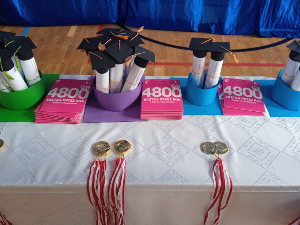 Gala Kornele 2023. Zdjęcie przedstawia stół nakryty białym obrusem, na którym leżą medale dla uczniów  i listy gratulacyjne dla  rodziców. 