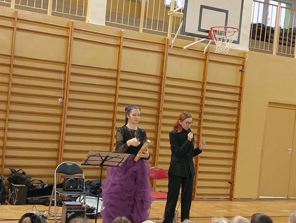 Koncert filharmonii. Na zdjęciu stoją dwie panie. Jedna trzyma w rękach elementy drewniane skrzypiec, druga opowiada o historii powstania skrzypiec. 